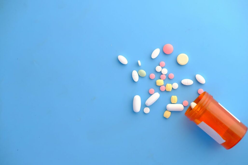 An orange prescription pill bottle tipped over on a solid blue background, spilling a variety of white, pink, and yellow pills.