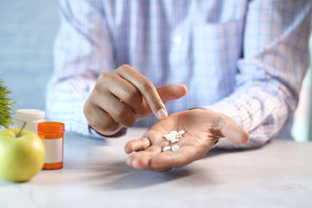 A close-up of a person holding several small white pills in their open palm, with an orange pill bottle and a green apple nearby.