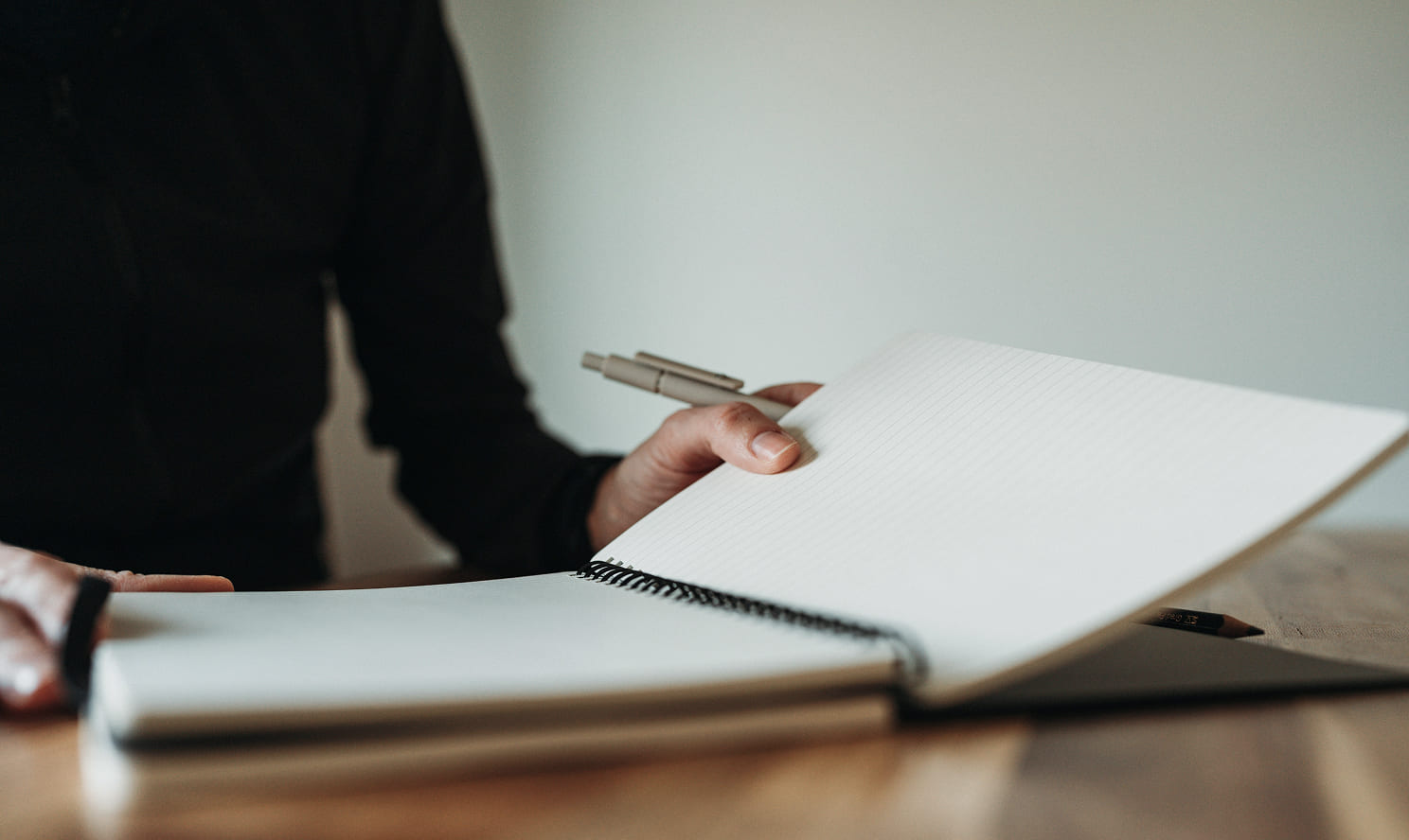 Close-up of hands holding an open, lined spiral notebook and a pen at a wooden desk.