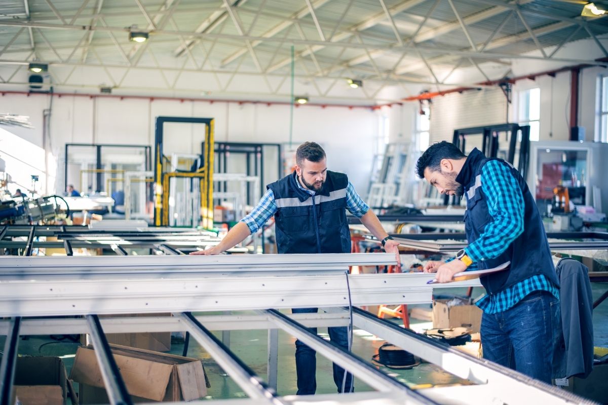 Two manufacturing workers inspecting and writing notes on a metal frame assembly on a factory floor.