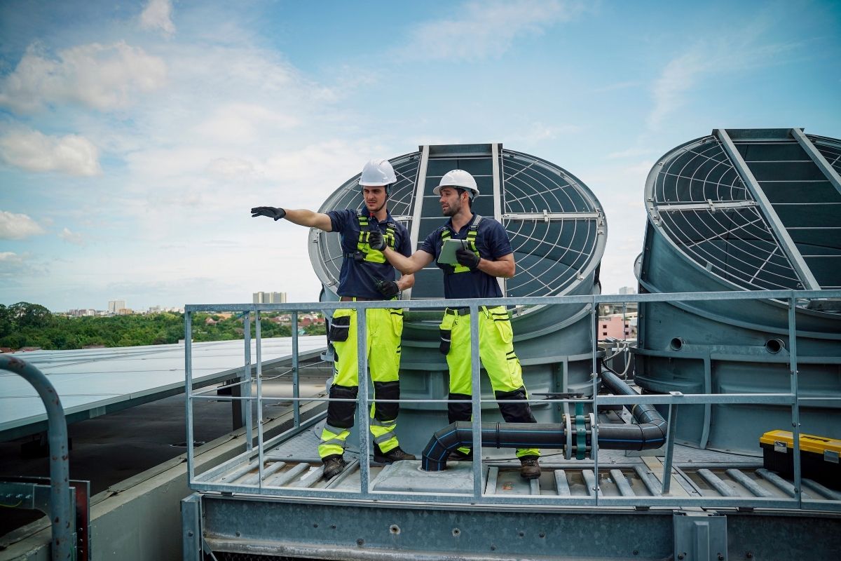 Two technicians in safety gear and hard hats inspecting large industrial HVAC cooling towers on a commercial rooftop, with one worker holding a tablet and pointing into the distance.